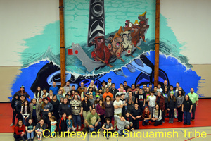 Photograph of women, young adults and children from the Suquamish Tribe in modern clothing, standing in front of a large mural wall that depicts bears rowing a canoe on top of a killer whale. The art style of the mural is typical of Pacific Northwest Native artwork. 