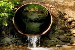 Photograph of water trickling out of a large metal pipe into a river. The pipe is embedded within a hill of dirt and the view of the photo is looking into and through the pipe.
